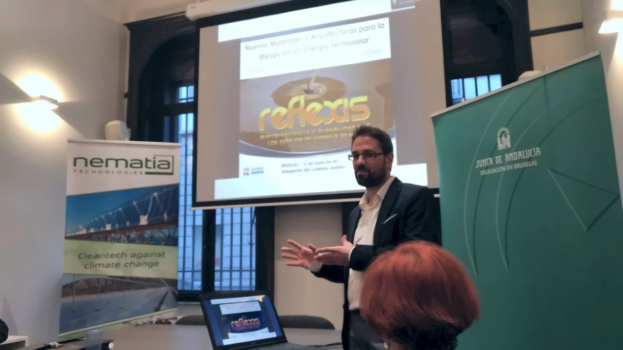 A speaker presents REFLEXIS in front of a screen, between the Nematia and Junta de Andalucía roll-ups