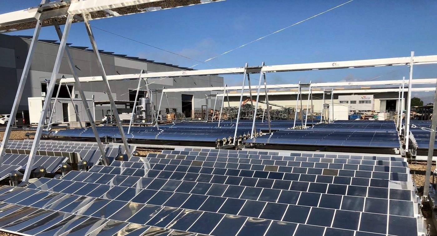 Rows of installed mirrors under the Fresnel collector structure, with industrial buildings and blue sky in the background