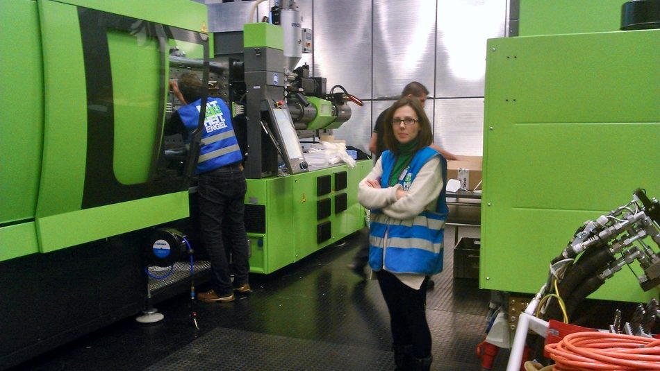 Test hall with green injection moulding machines and two people in visitor vests, one of them Zayda Donaire