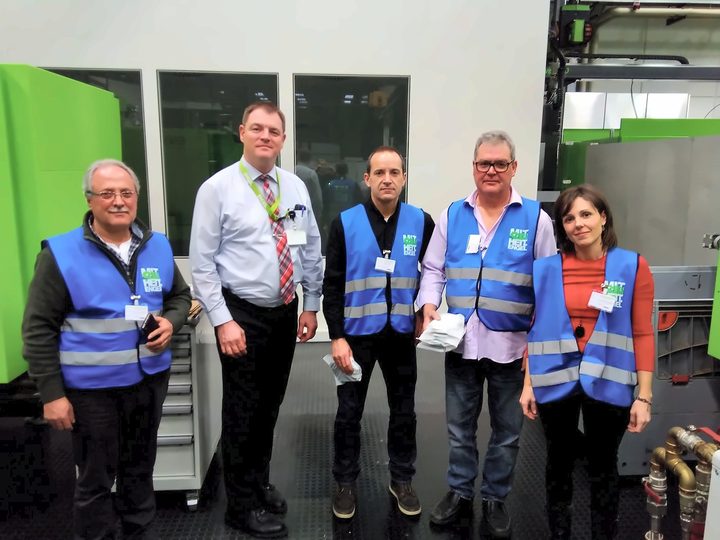 Five people in visitor vests pose next to a green injection moulding machine
