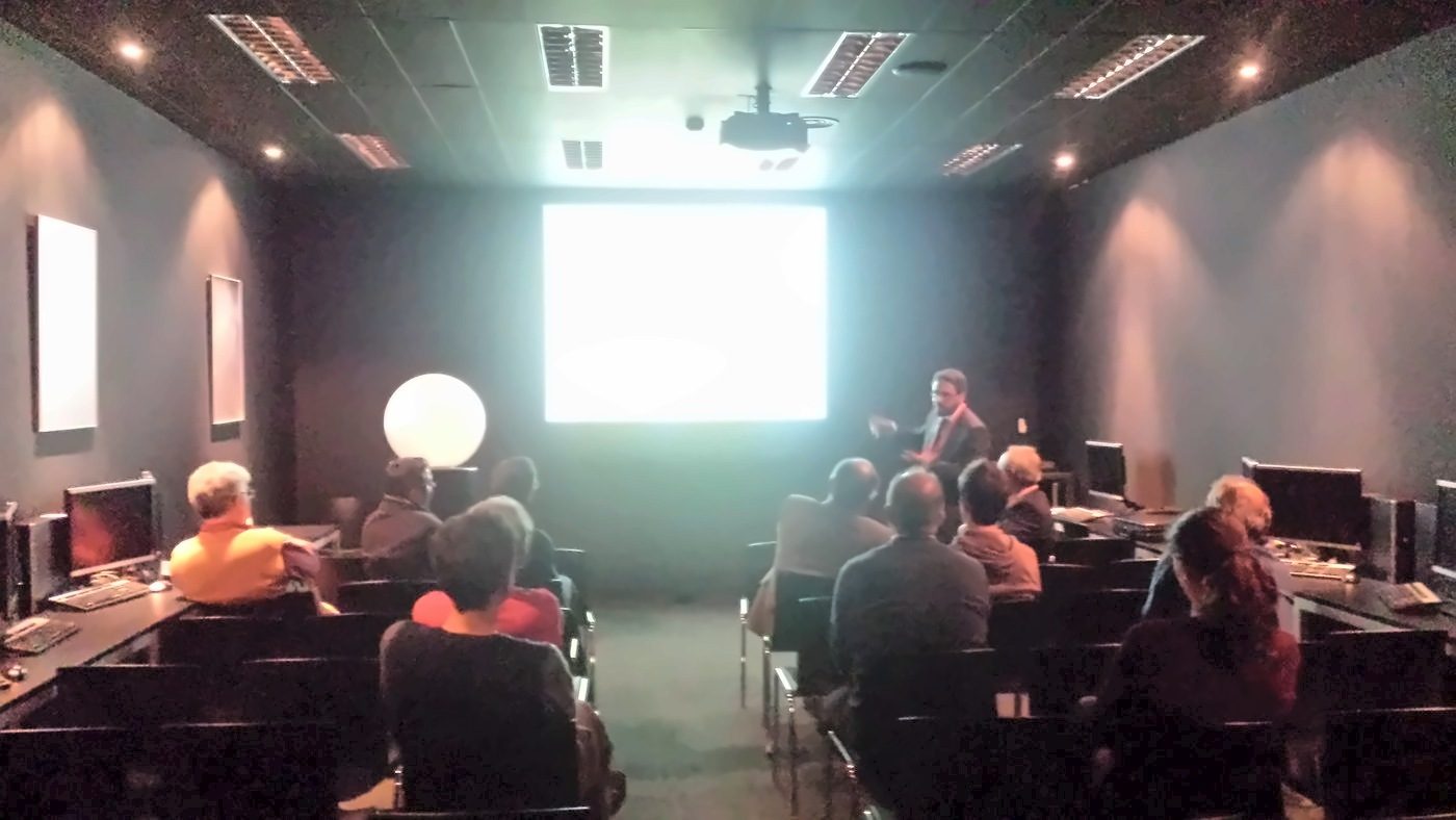 Darkened room with an audience in front of a lit screen, during a talk