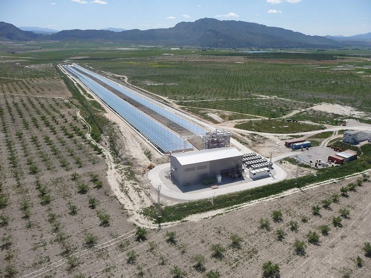 Aerial view of a Fresnel plant: a long row of flat mirrors under its raised receiver, next to the turbine building