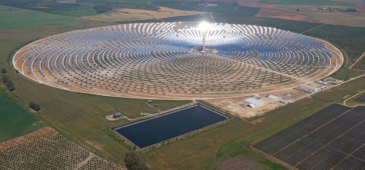 Aerial view of a circular field of heliostats concentrating light on a central tower, among farmland