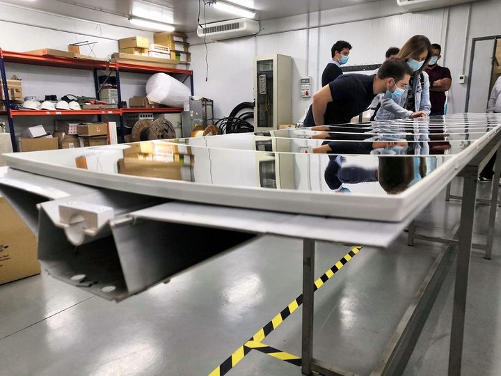 Several people in face masks inspect a long row of mirrors mounted on profiles in an industrial building